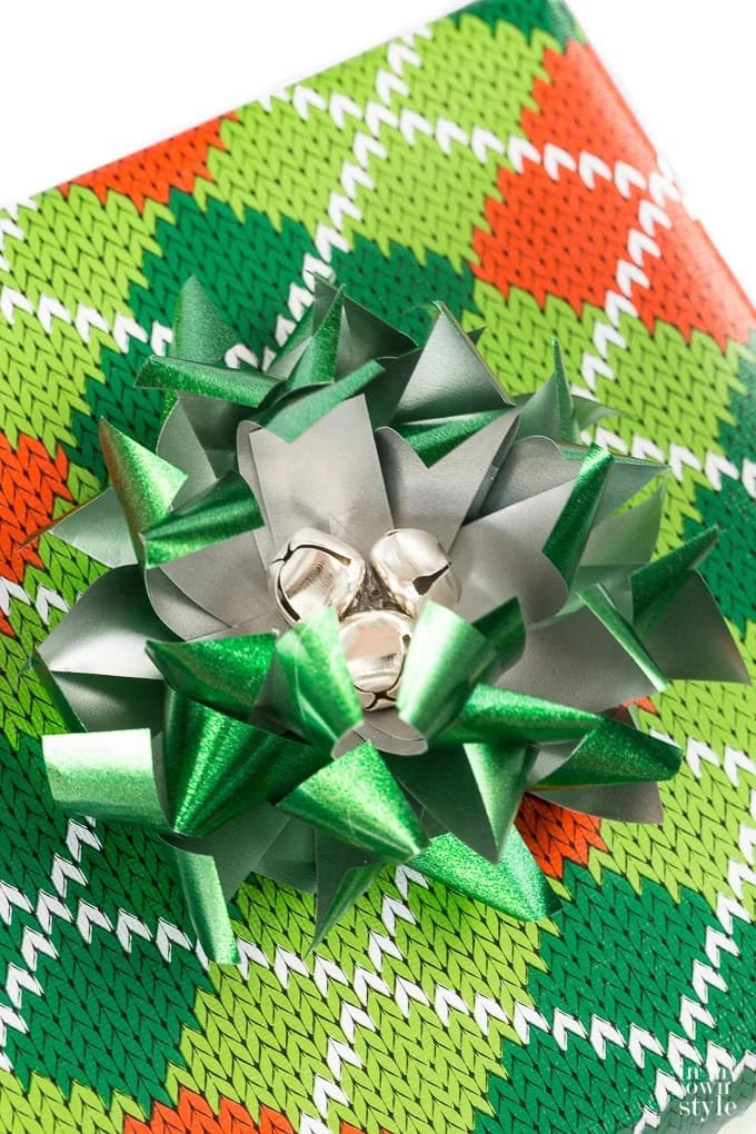 A vibrant green, orange, and white gift wrap with a shiny green bow and bells, presenting a beautifully unique gift wrapping idea with a holiday sweater theme.