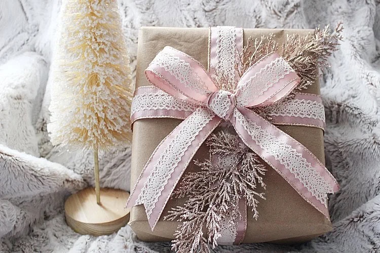 A gift wrapped in kraft paper with a blush lace ribbon and frosted branch, exemplifying beautifully unique gift wrapping ideas with a soft, elegant touch.