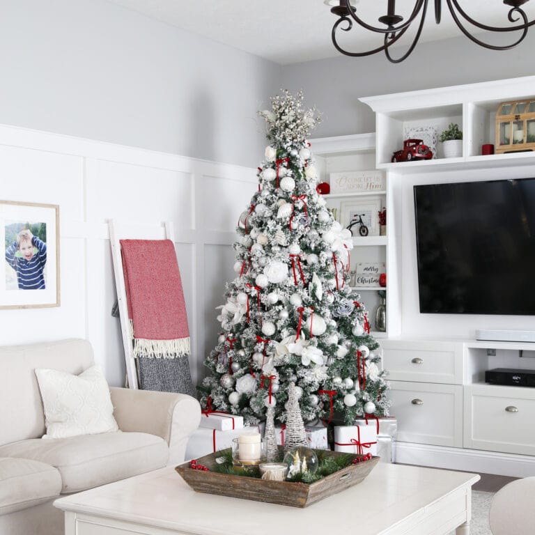 A beautifully decorated living room showcases a snowy Christmas tree adorned with red and white ornaments, embodying expert tips on how to decorate a Christmas tree. The room features a cozy beige sofa, a wall-mounted TV, a festive centerpiece on the coffee table, and a photo ladder with blankets.