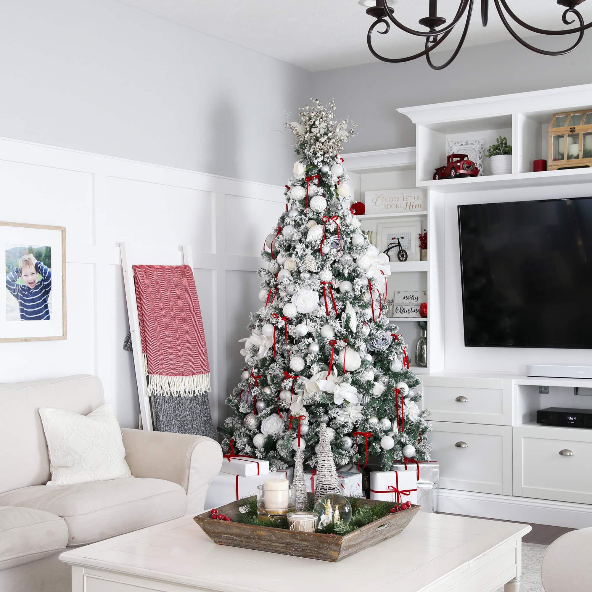 A beautifully decorated living room showcases a snowy Christmas tree adorned with red and white ornaments, embodying expert tips on how to decorate a Christmas tree. The room features a cozy beige sofa, a wall-mounted TV, a festive centerpiece on the coffee table, and a photo ladder with blankets.